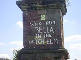 Photograph of graffiti reading “Who Put Bella in the Wych Elm?” written on a brick pillar near Hagley Wood, referencing Britain’s enduring unsolved wartime mystery.
